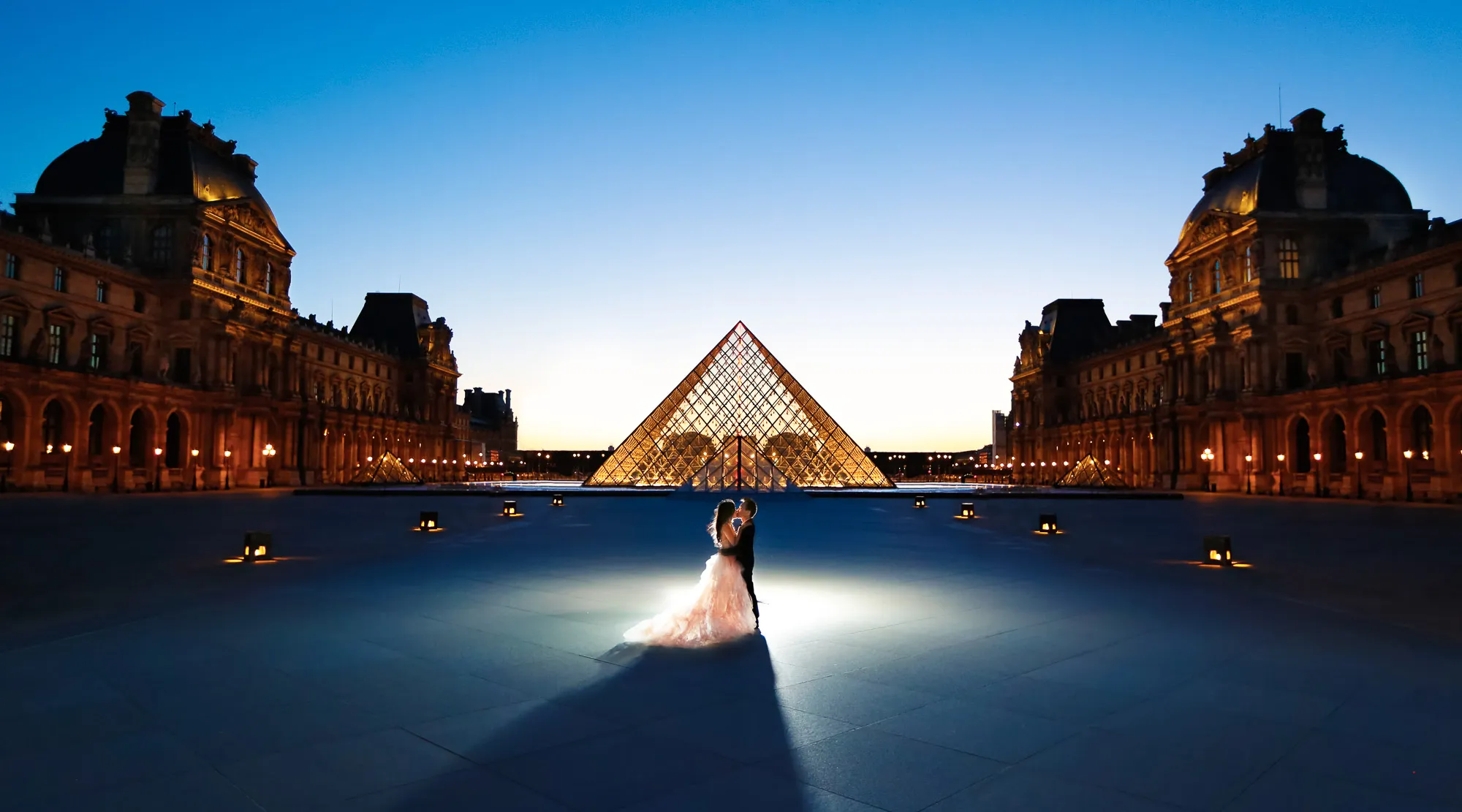 Romantic couple embracing at the Louvre Pyramid in Paris at dusk. Grand Louvre buildings glow under a deep blue sky.