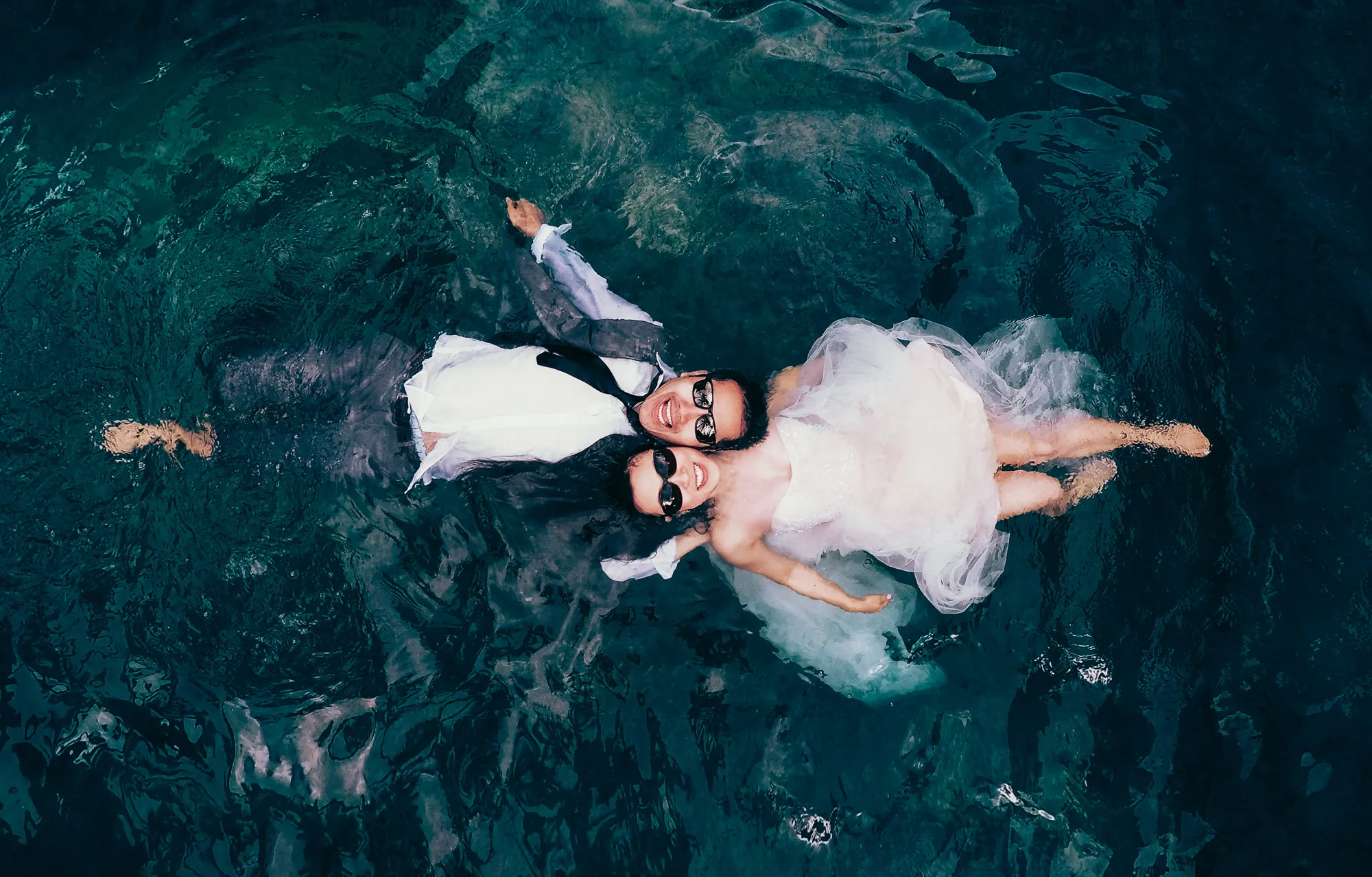 Joyful bride and groom in wedding attire and sunglasses float on their backs in clear, dark water, smiling.