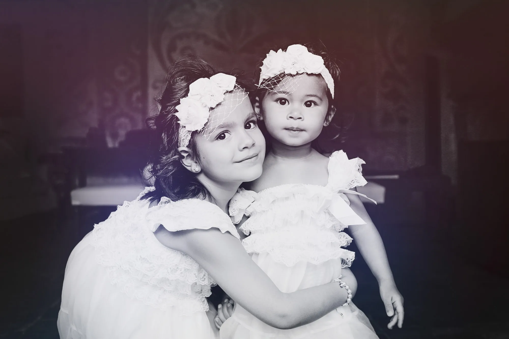 Two young girls in white lace dresses and floral headbands embrace, smiling sweetly. SixSixty Studio.