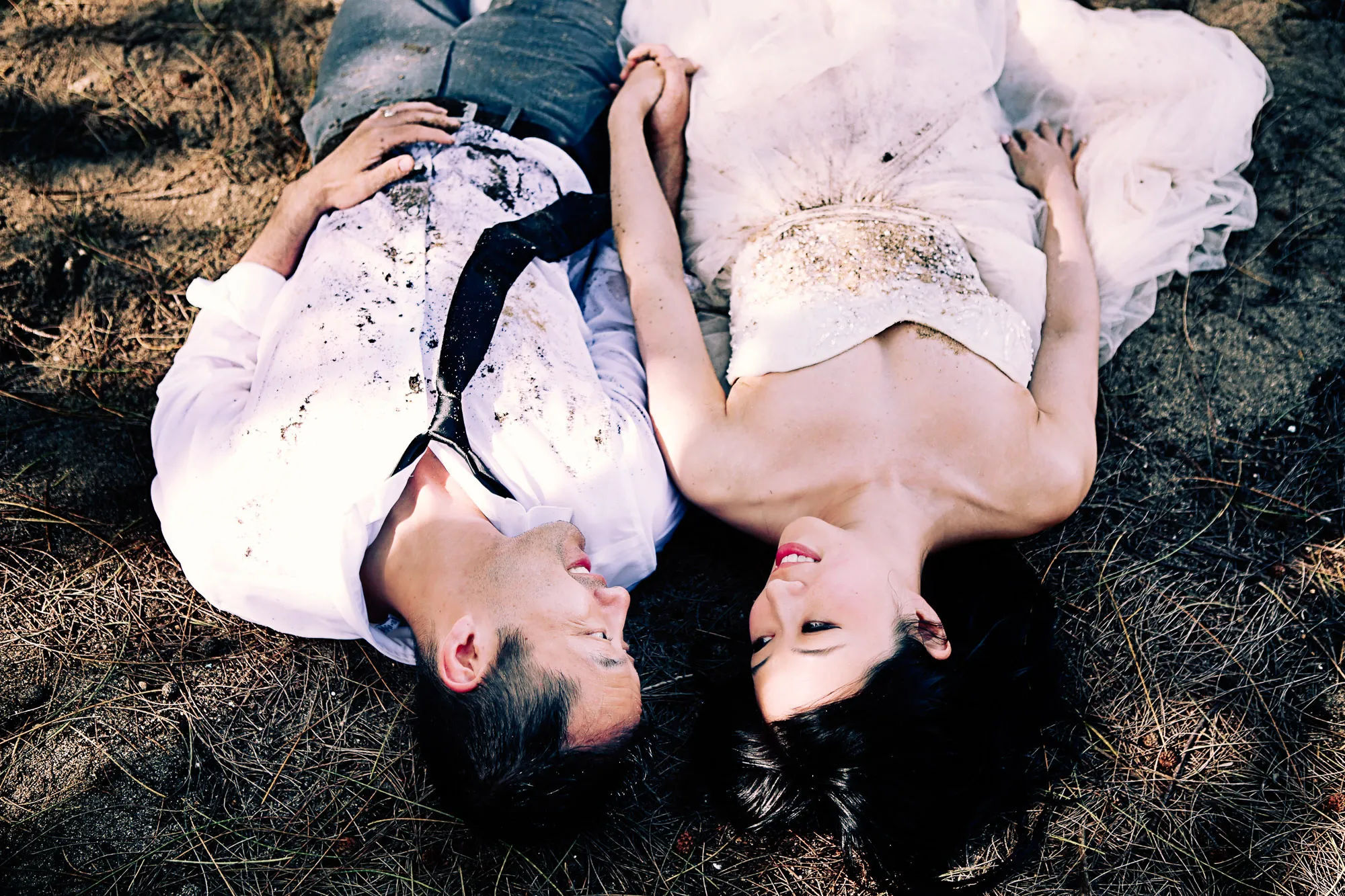 Bride and groom lie on sandy ground, holding hands, looking at each other. Romantic and playful outdoor portrait.