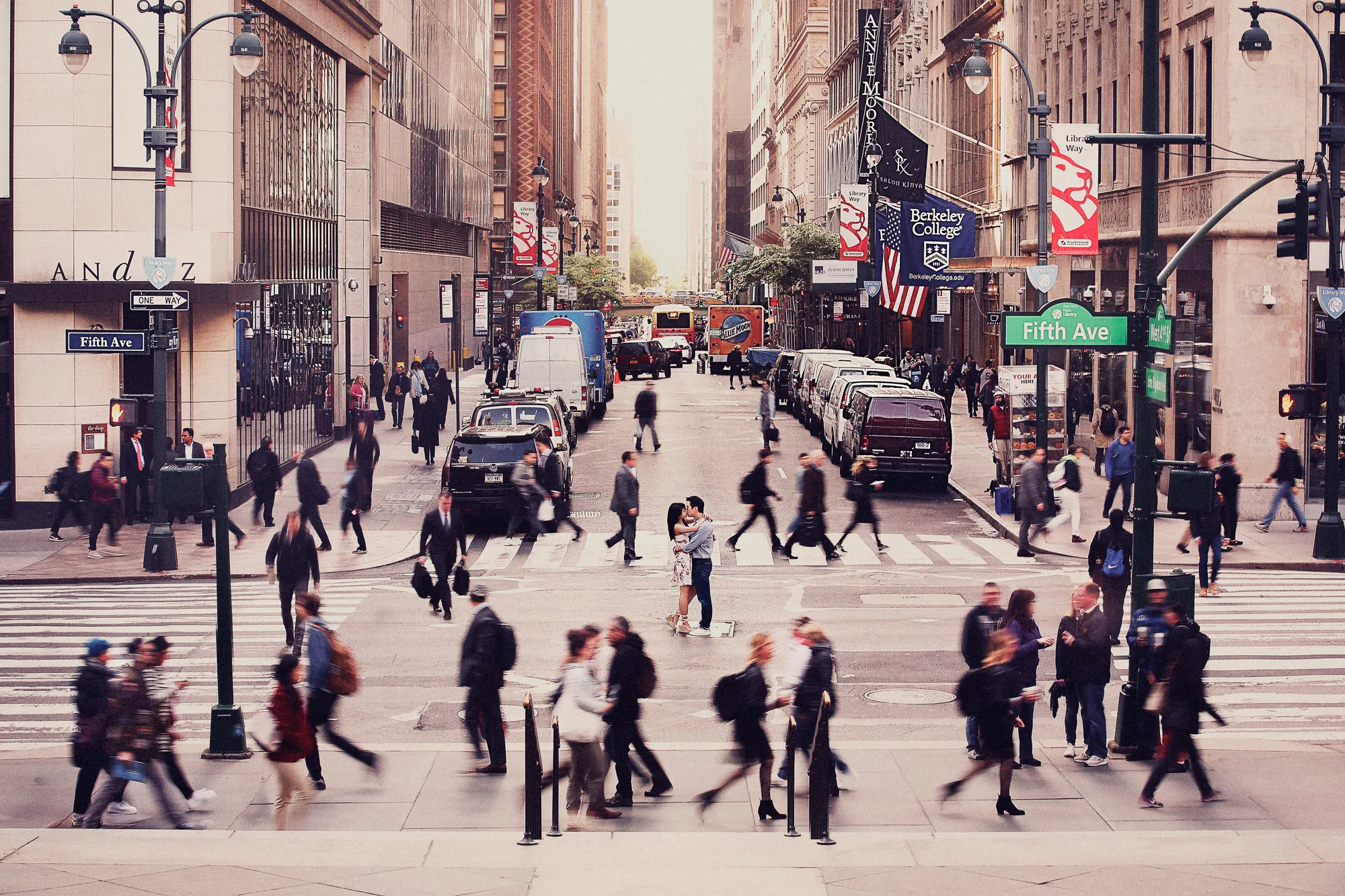 Bustling Fifth Ave street scene with blurred pedestrians and vehicles. A couple shares a kiss, a still moment in the city's rush.