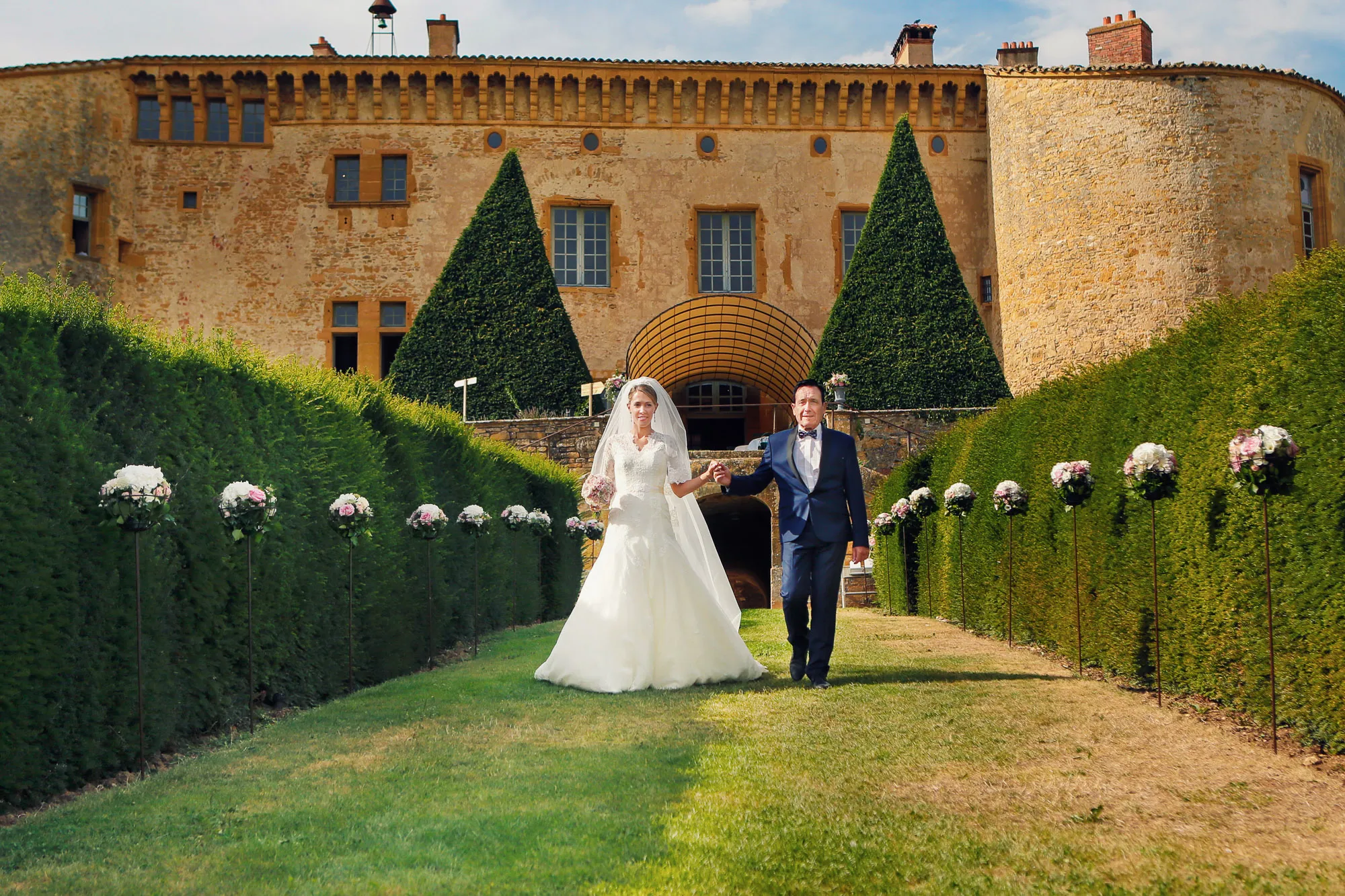 Joyful bride and groom walk hand-in-hand on a garden path lined with flowers, in front of a grand stone castle.