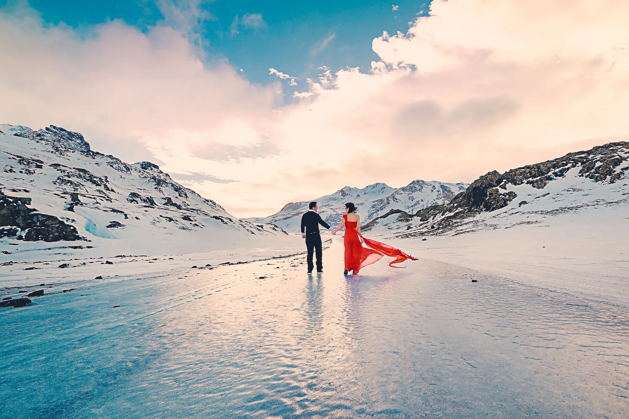 Couple in red dress and suit walk hand-in-hand on frozen lake amidst snowy mountains under dramatic sky. Romantic.