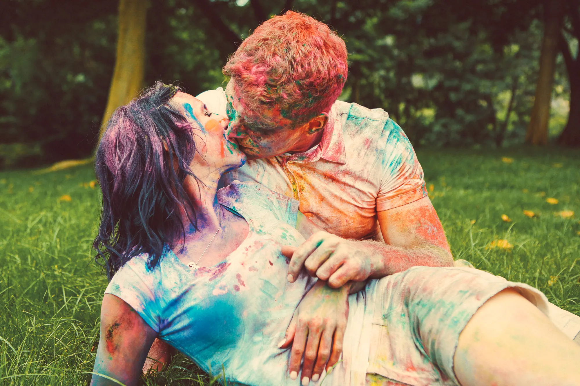 A couple covered in vibrant powder kisses passionately on green grass.