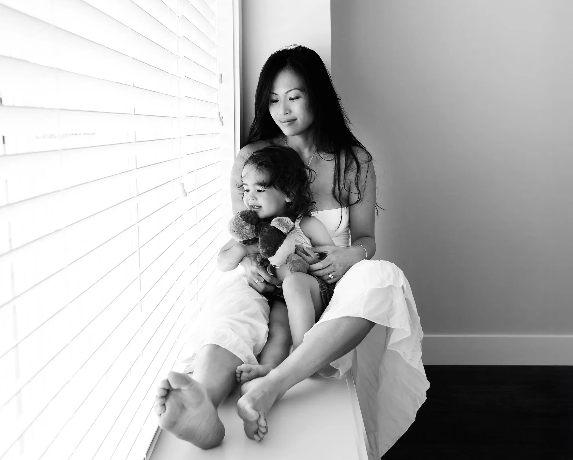 B&W photo: Mother and child share a tender moment on a window sill. Child holds a toy and looks out.