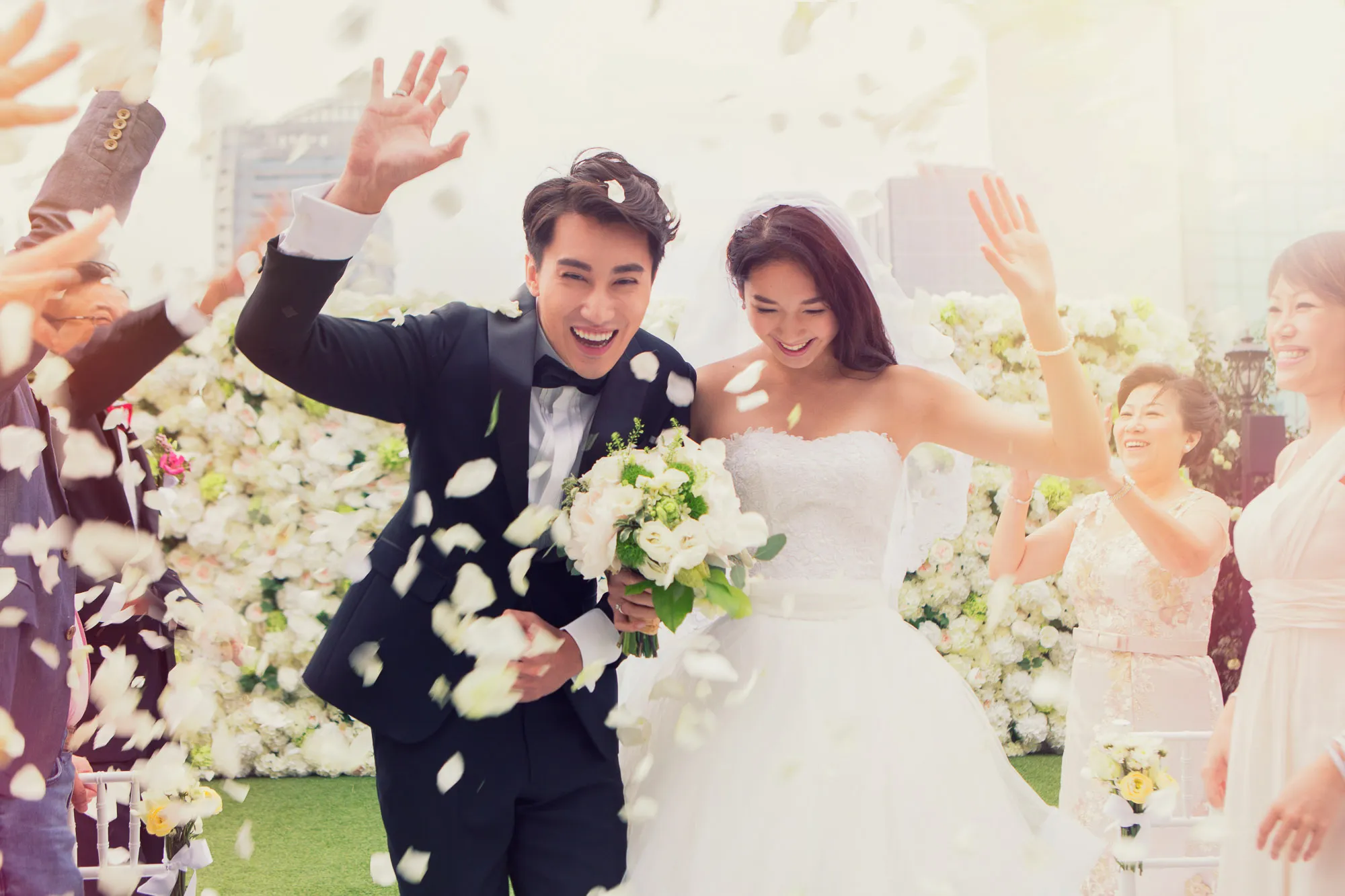 Joyful bride & groom with guests at an outdoor wedding, showered in petals. Flower wall backdrop. SixSixty Studio.