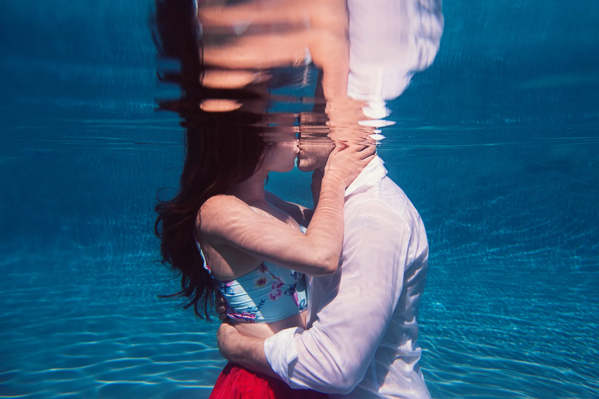 Romantic couple kissing underwater in a pool, with reflections on the surface.