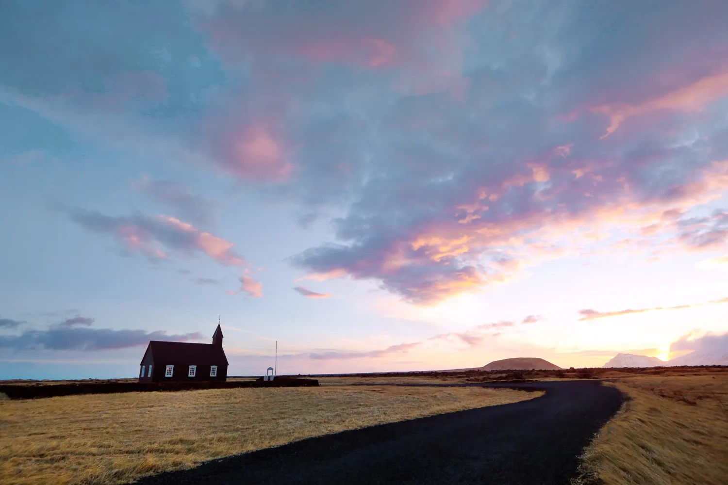 Red wooden church with white windows stands alone in barren landscape at sunset.