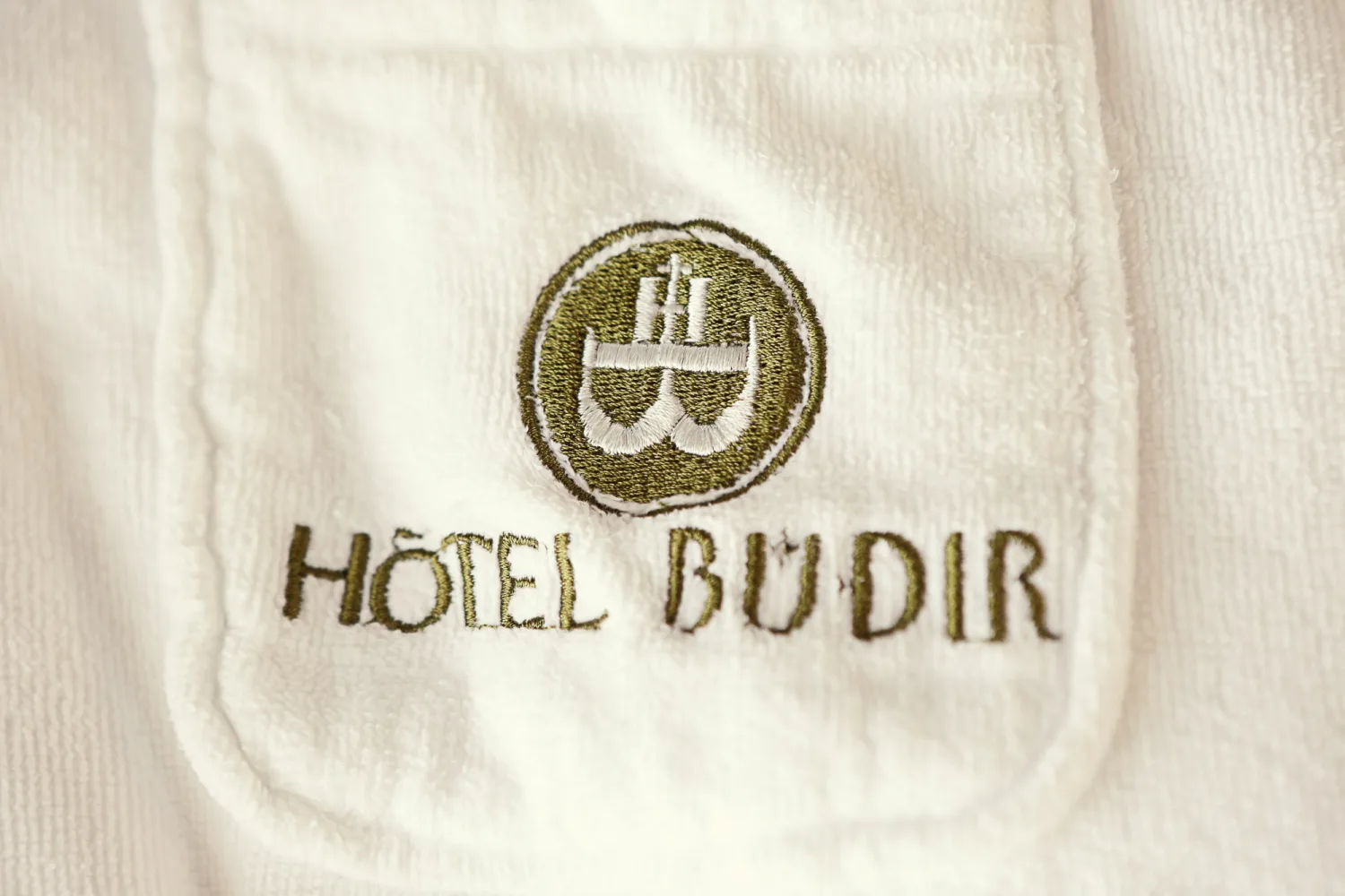 Embroidered Hotel Budir logo on white linen fabric with olive-green thread.
