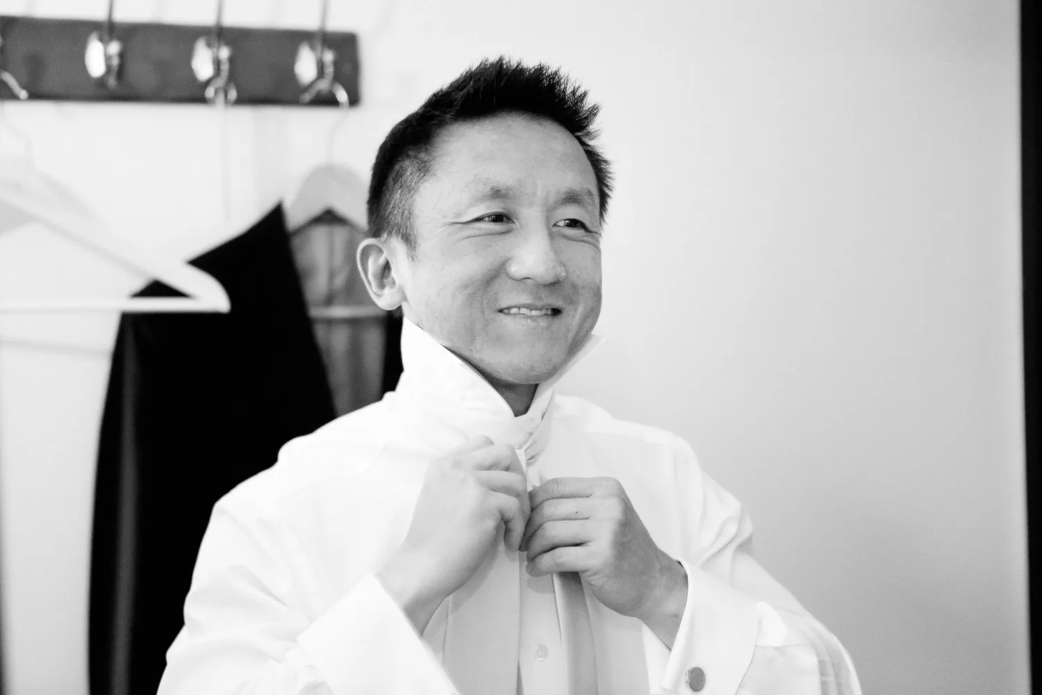 Man in white dress shirt buttoning up in indoor dressing room with mirror lights.