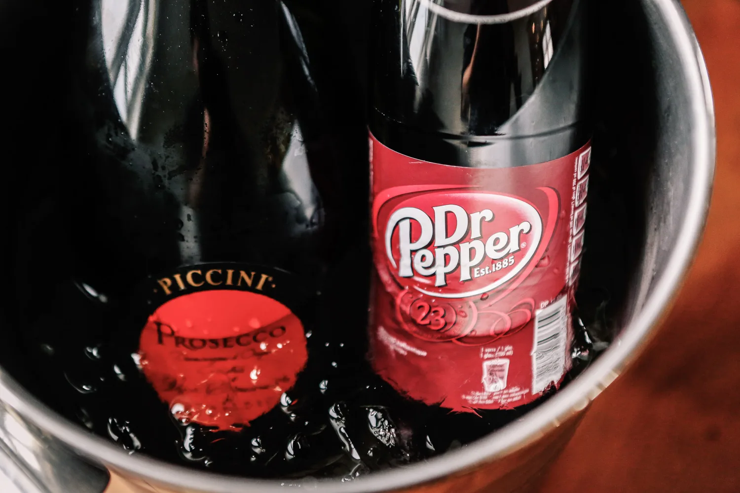 Dr Pepper and Prosecco bottles chilling in ice bucket with red-orange background.