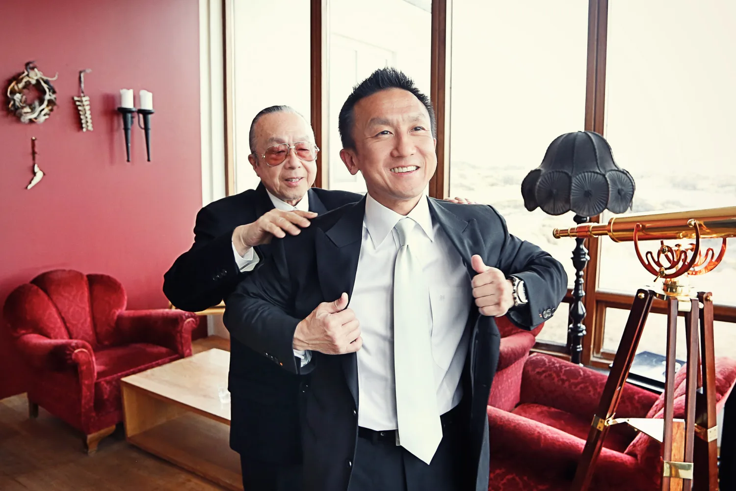 Two men in dark suits and ties pose in a pink-walled room with red velvet furniture.