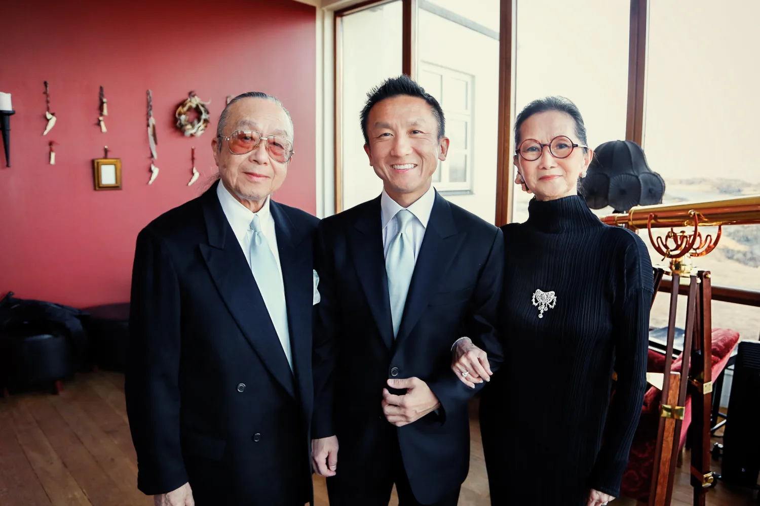 Three people in formal dark suits pose indoors against a pink wall with decorative items.