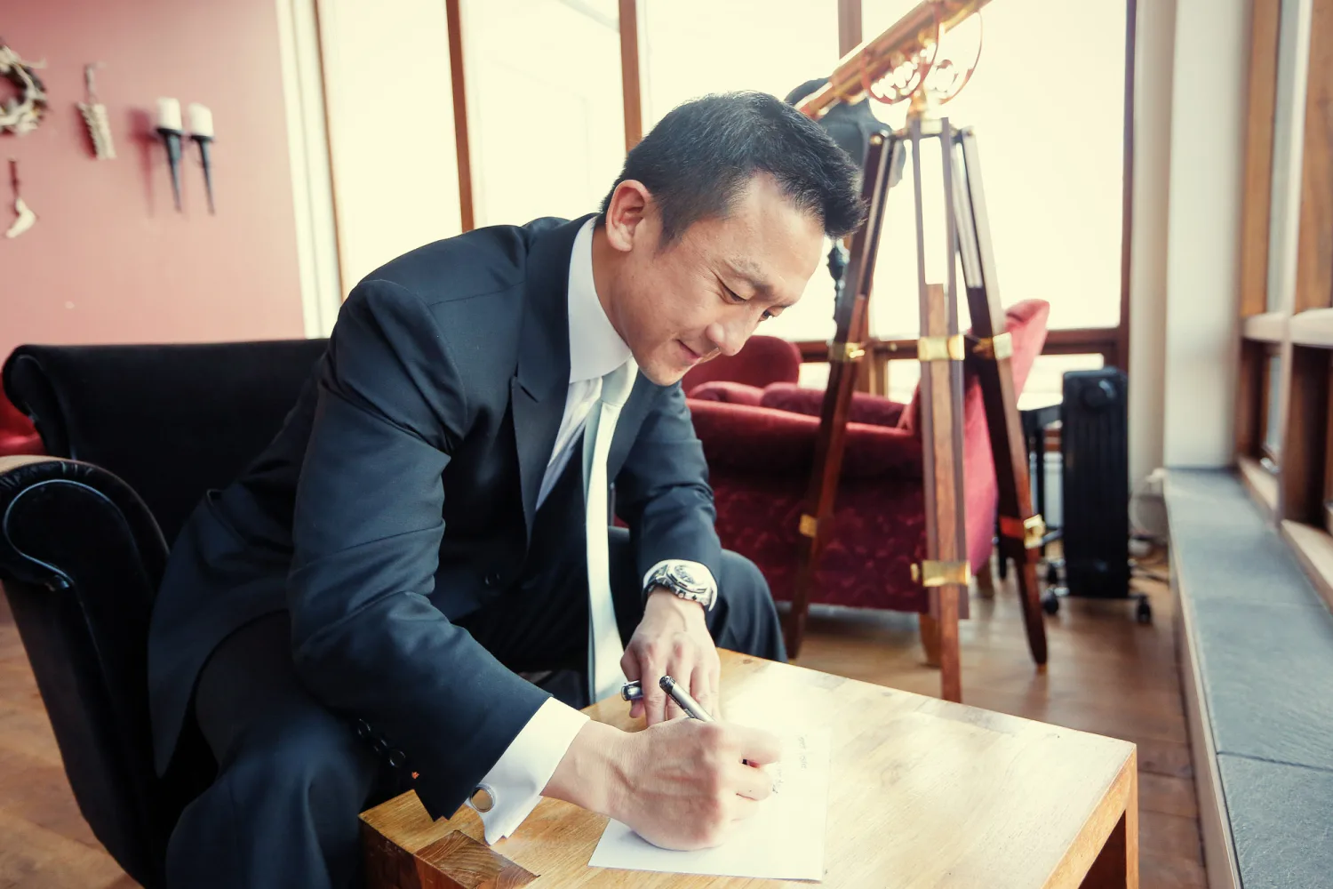 Man in dark suit and white shirt signing document at wooden table indoors.