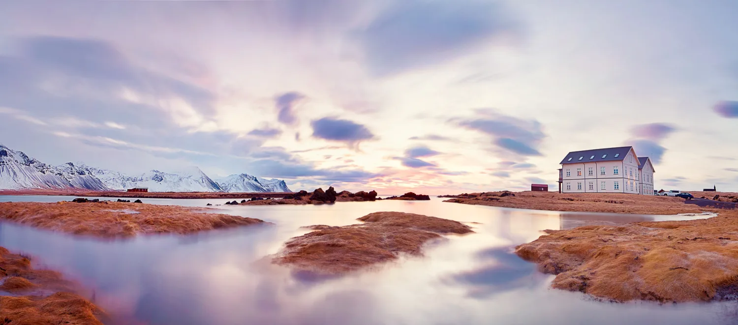 Pink house on Arctic coastline at dusk with snow-capped mountains and tidal pools.