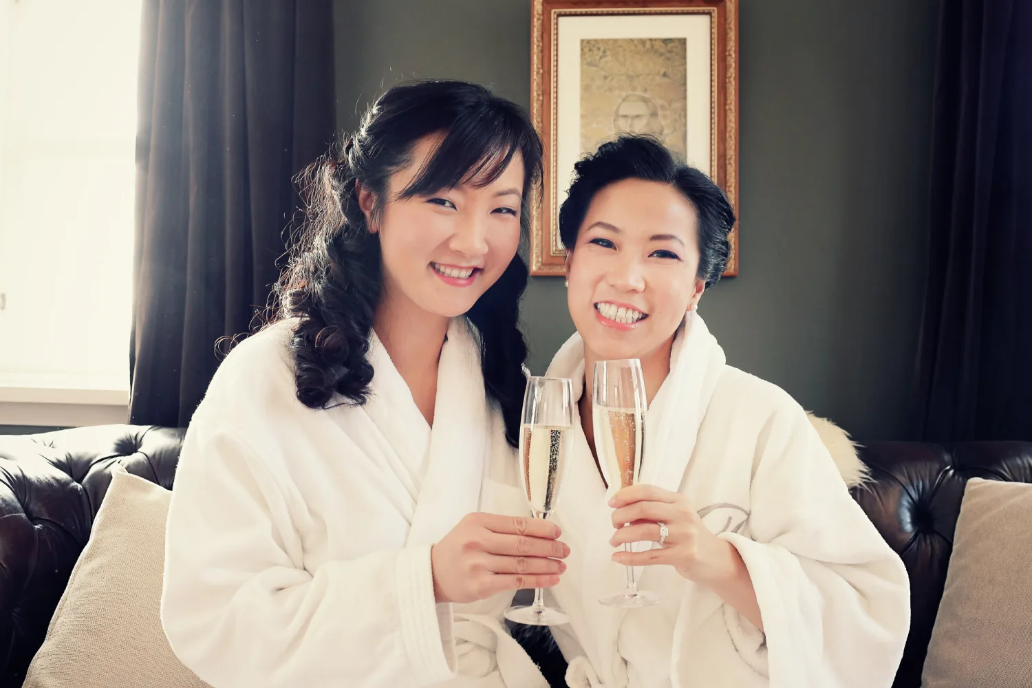 Two people in white robes holding champagne glasses indoors on a black sofa.