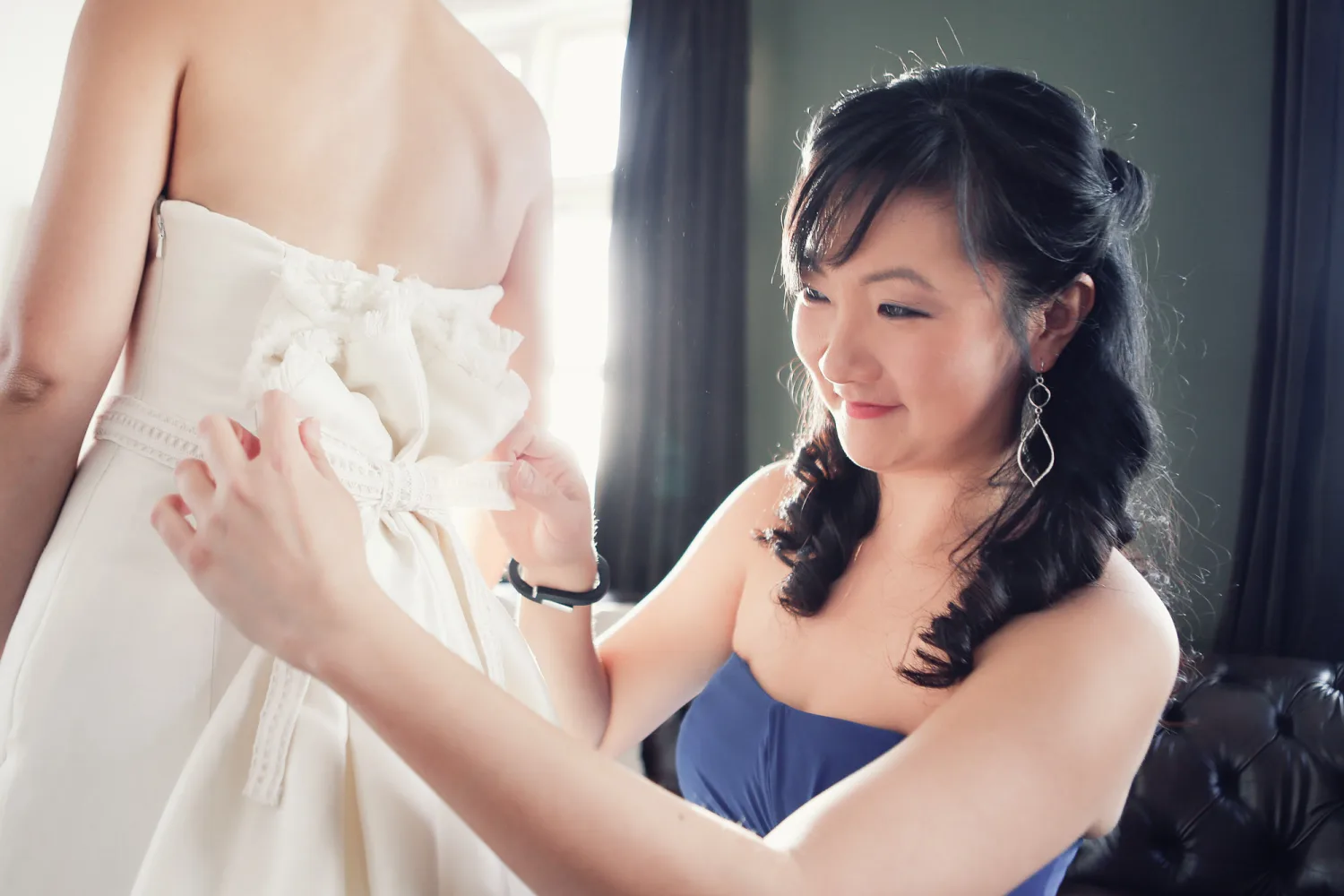 Woman in blue dress adjusts white wedding dress bow on bride indoors.