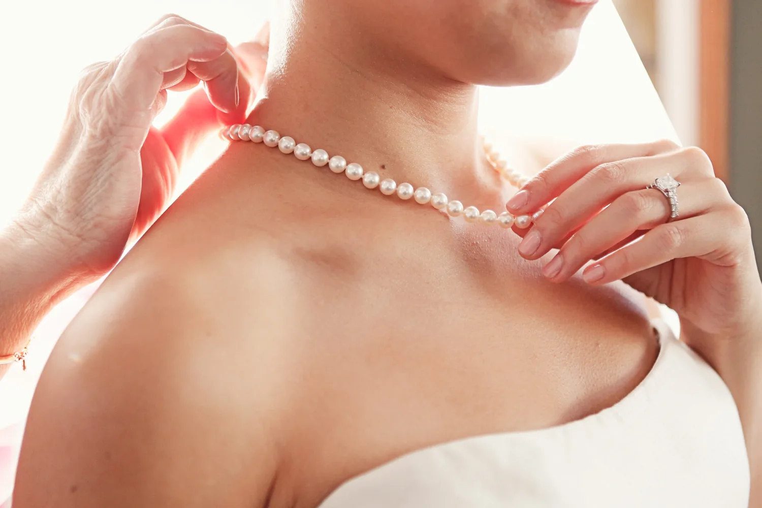 Bride fastening pearl necklace while wearing diamond engagement ring and white dress.