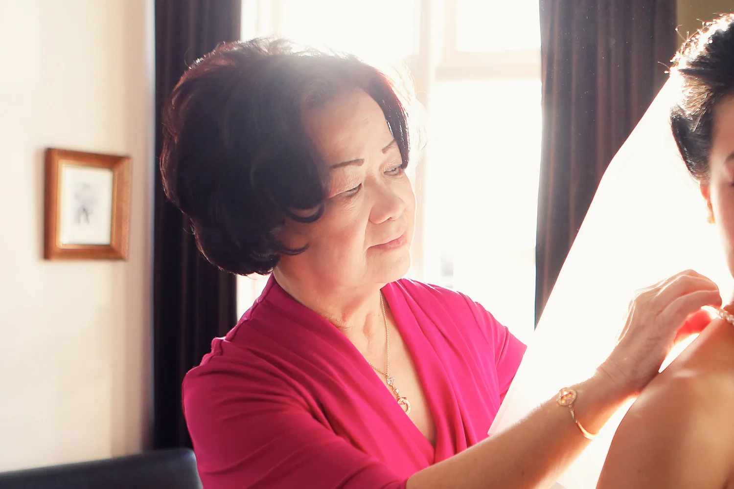 Woman in bright pink top adjusts another person's clothing by a sunlit window.