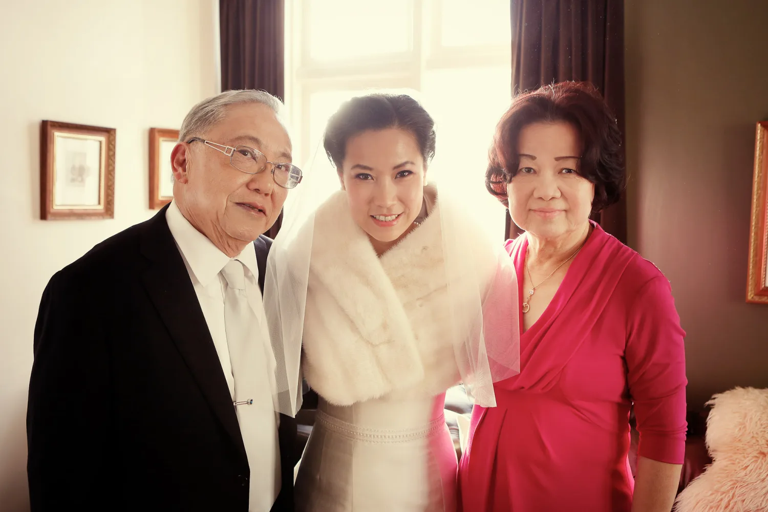 Bride in white dress with veil poses with two guests in formal attire indoors.