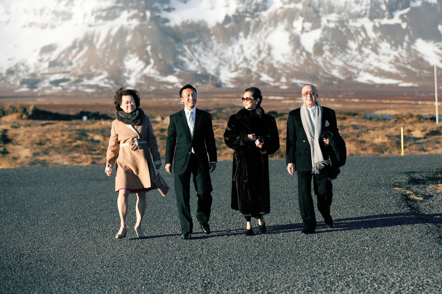 Four people in formal attire walking on a road with snow-capped mountains in the background.