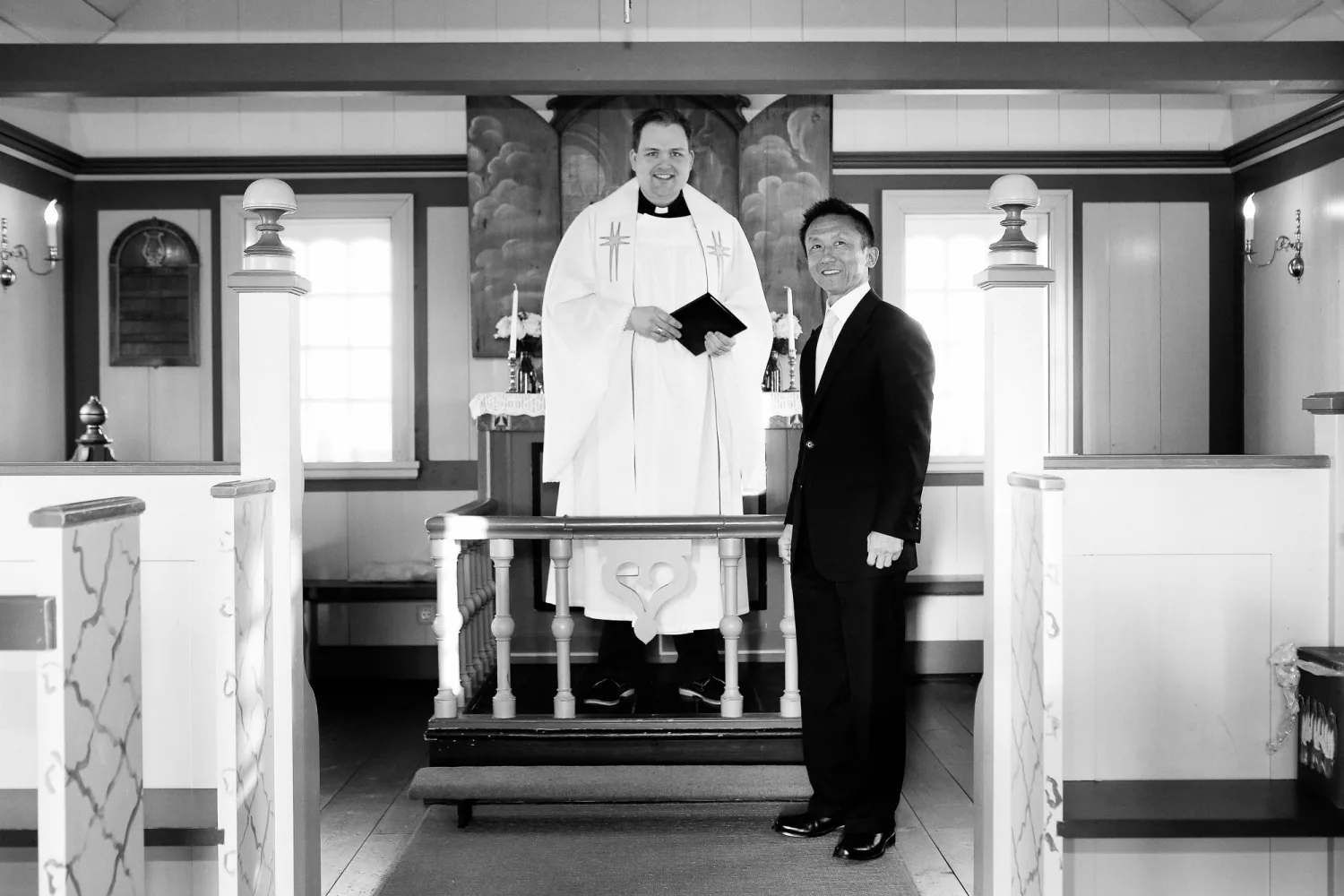 Clergy member in white vestments holds prayer book at altar beside man in black suit in church interior.