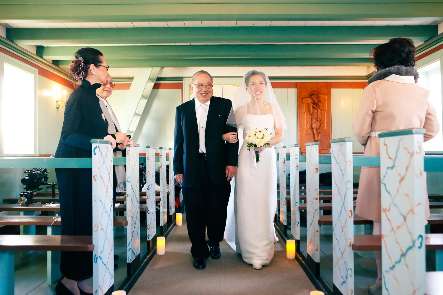 Bride in white gown and veil walks down church aisle with man in dark suit, flanked by decorated pews.