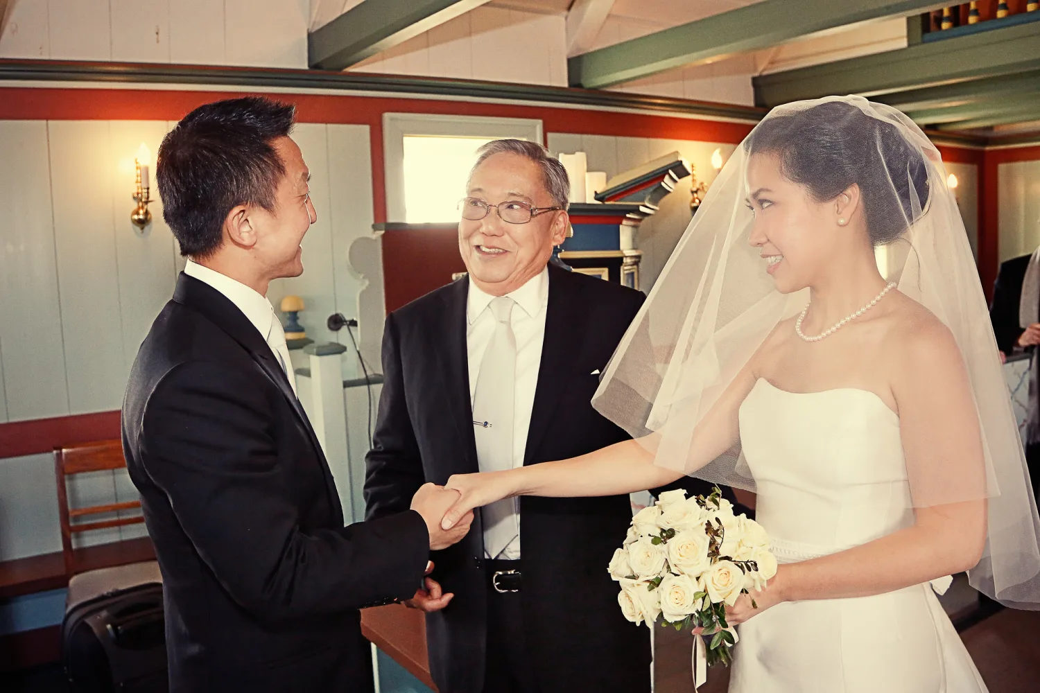 Bride in white strapless gown and veil shakes hands with groom in black tuxedo indoors.