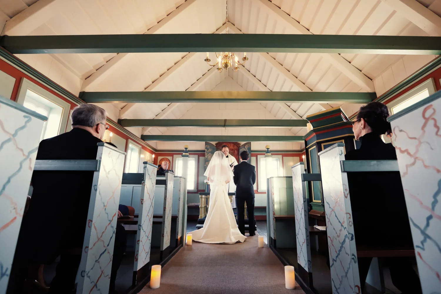 Bride in white gown and groom in black suit stand at altar inside chapel with wooden beams and stained glass windows.