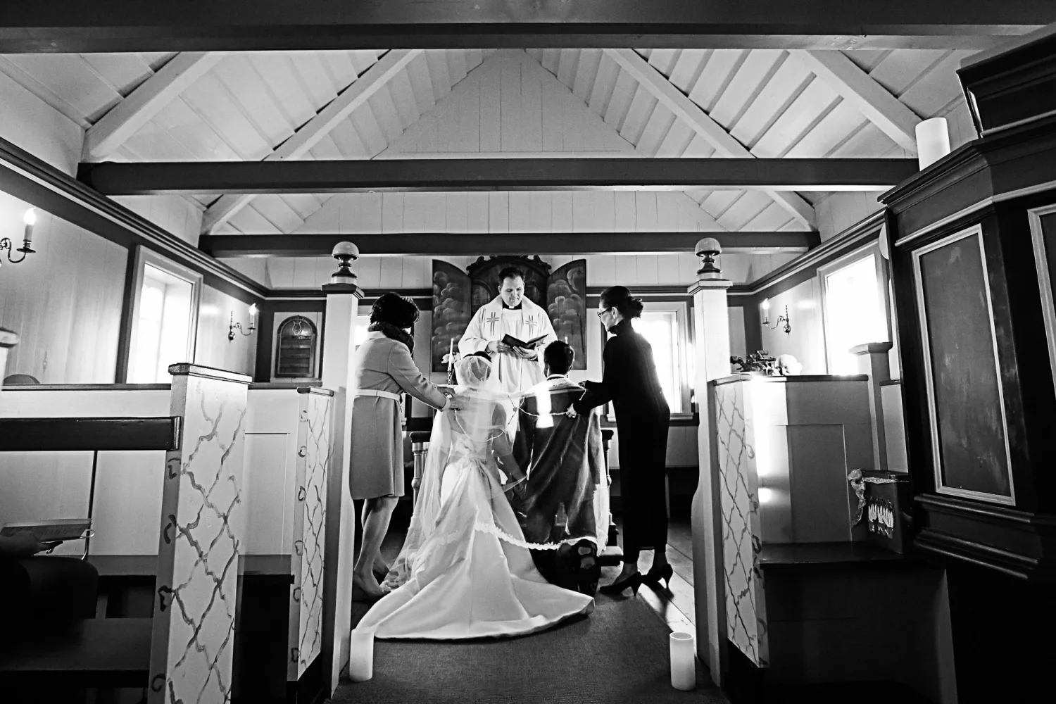 Bride in white gown with train stands before officiant in chapel during wedding ceremony.