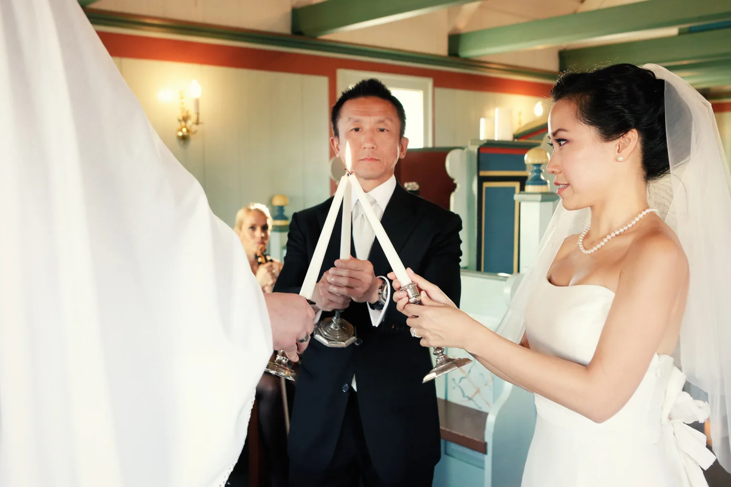 Bride in white strapless gown and veil holds candle with groom in black tuxedo during indoor ceremony.