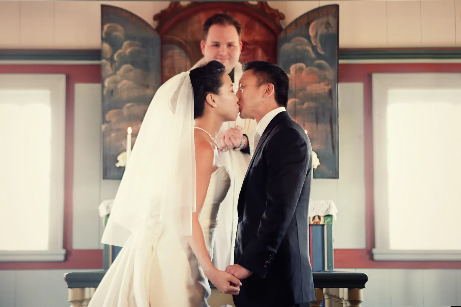 Bride in white veil and groom in dark suit kiss at altar in ornate chapel.