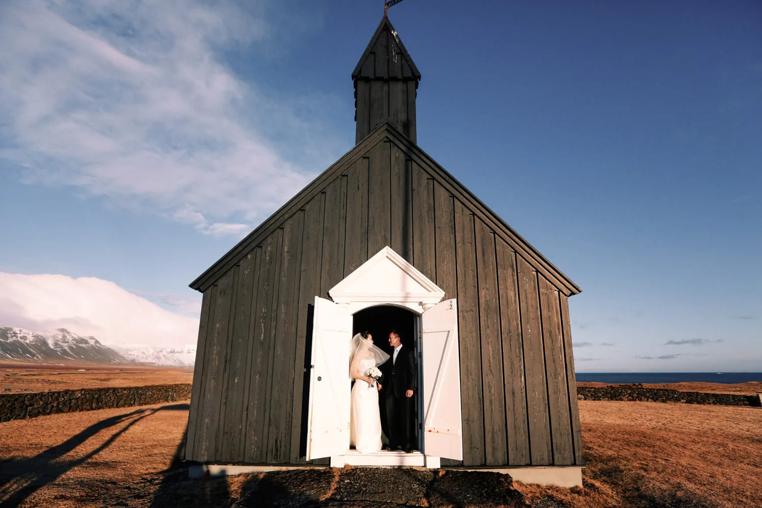 Two people in wedding attire stand in doorway of black wooden church with white entrance against snowy mountains and ocean.
