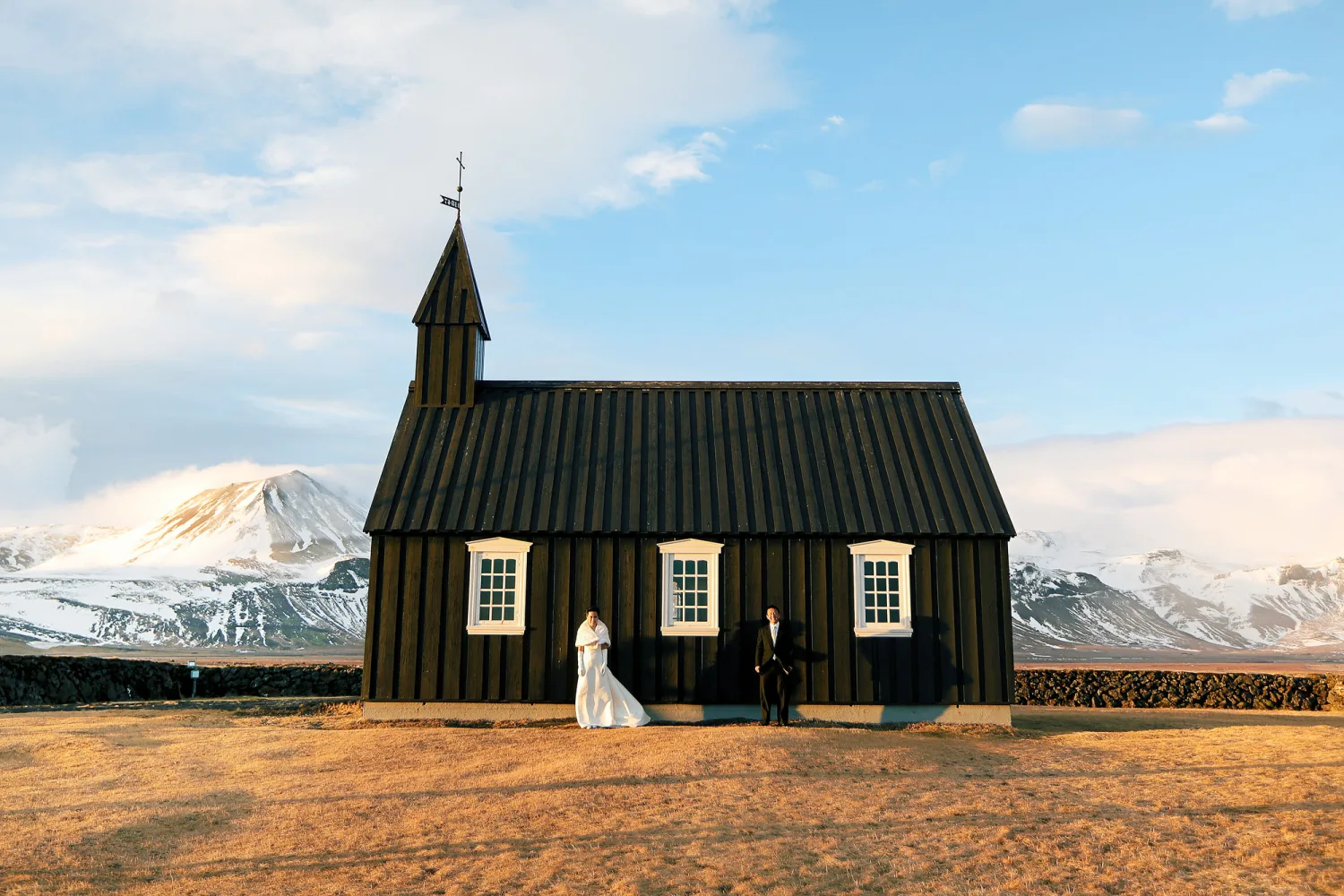 Two people in wedding attire stand before a black wooden church with white-trimmed windows against snow-capped mountains.