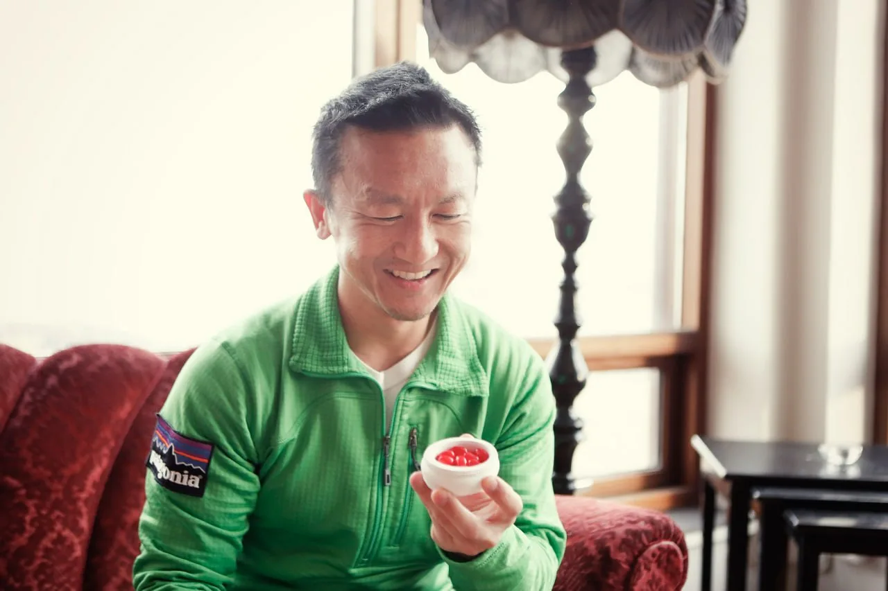 Smiling man in a bright green jacket holding a white bowl of red jelly beans indoors.