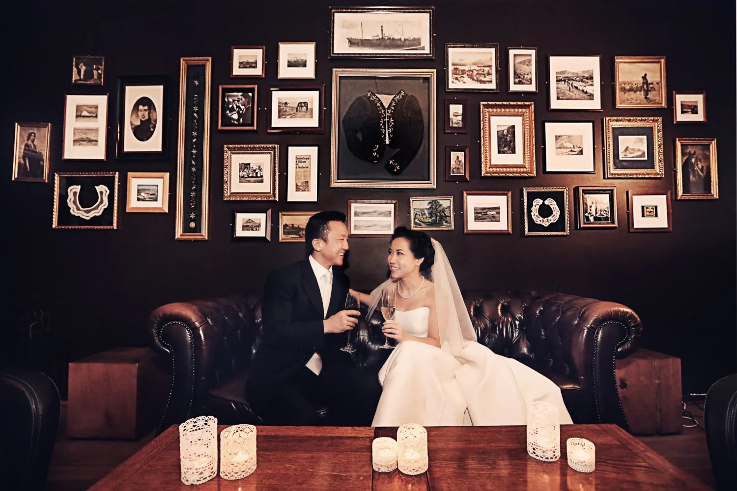 Bride and groom sit on leather sofa holding champagne glasses beneath gallery wall of framed photographs.