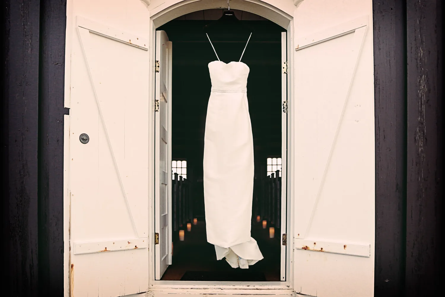 White strapless wedding dress hanging in doorway with dark interior and lit candles visible.
