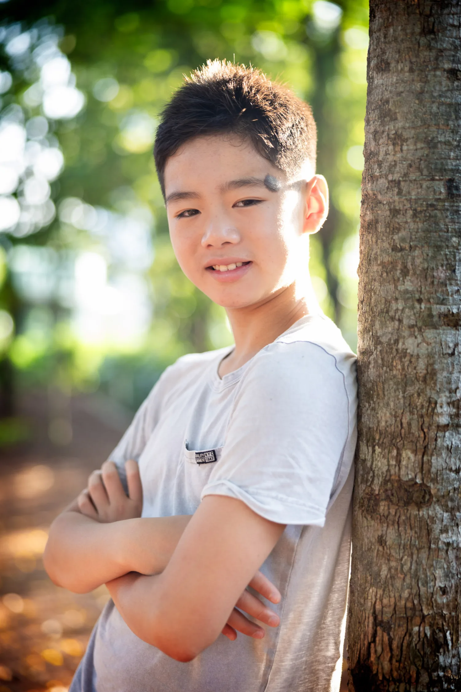 Avery leaning casually against a tree trunk, smiling softly in dappled sunlight