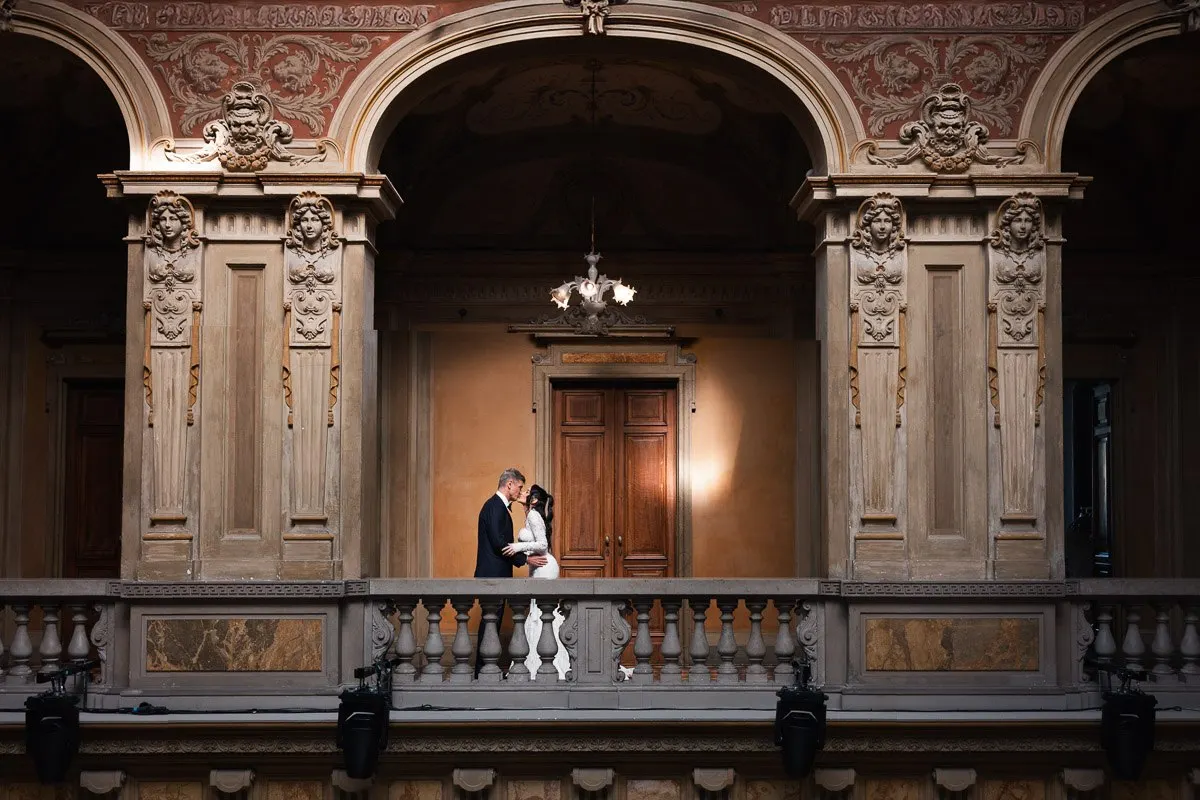 Vera and Emilio on Villa Erba's ornate balcony with their small white dog, warm light glowing through the arches behind them