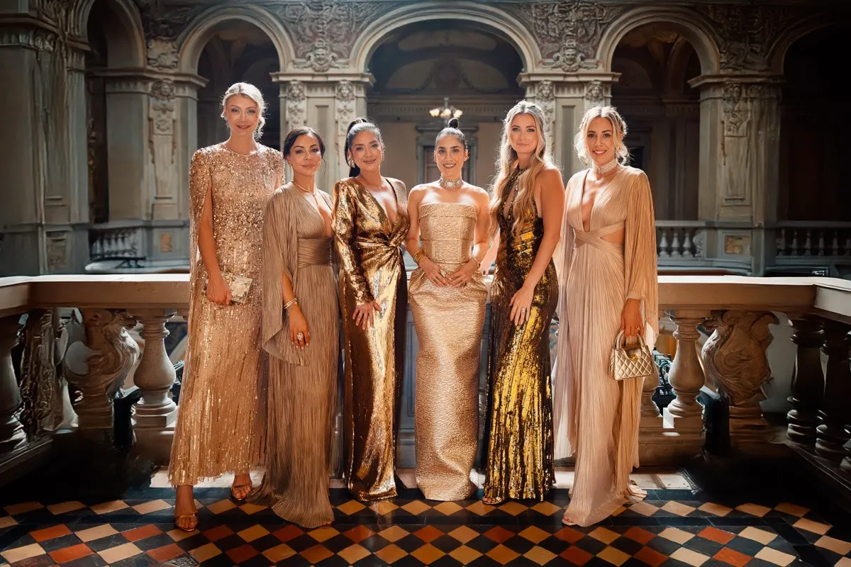 The bridesmaids in gold gowns lined up in Villa Erba's grand hall, ornate carved panels and a checkered floor behind them