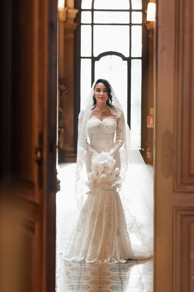 Vera framed in the villa's ornate doorway, natural light flooding through the window above her veil and lace gown