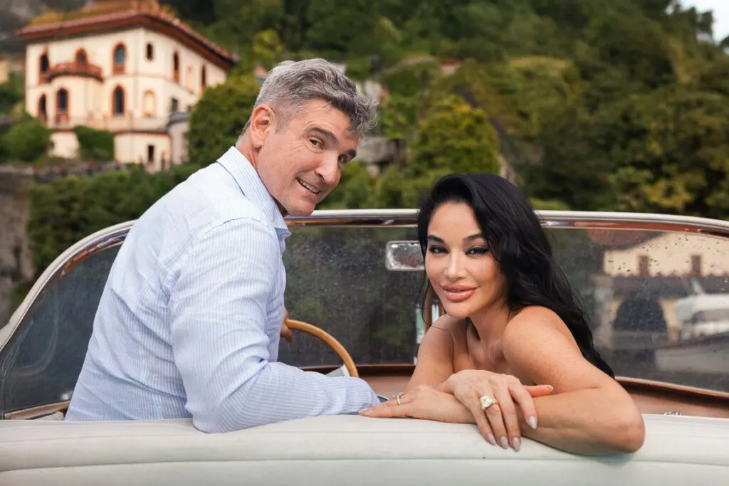 The couple in a vintage car, the pink facade of a lakeside villa glowing behind them