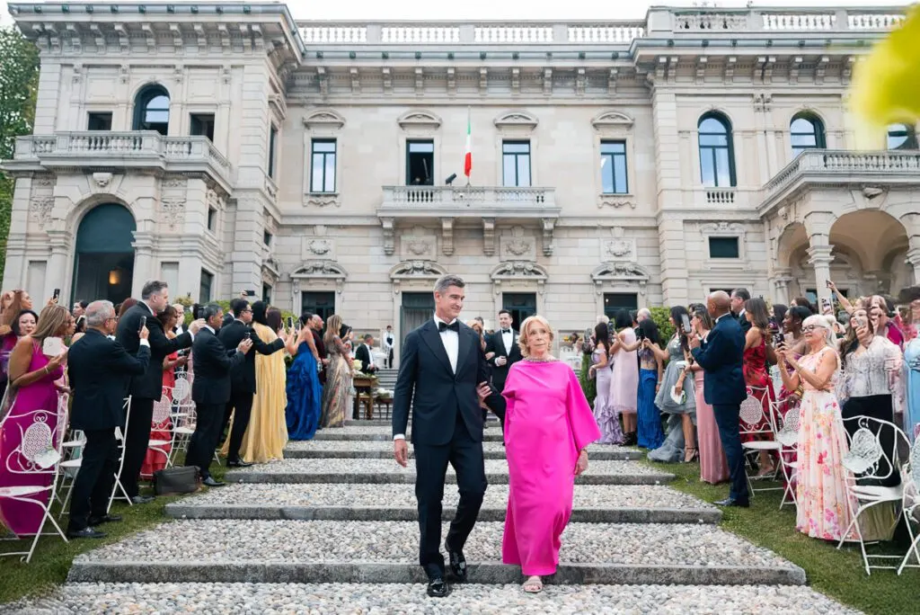 The groom walking down the aisle with his mother, guests filling the courtyard of Villa Erba