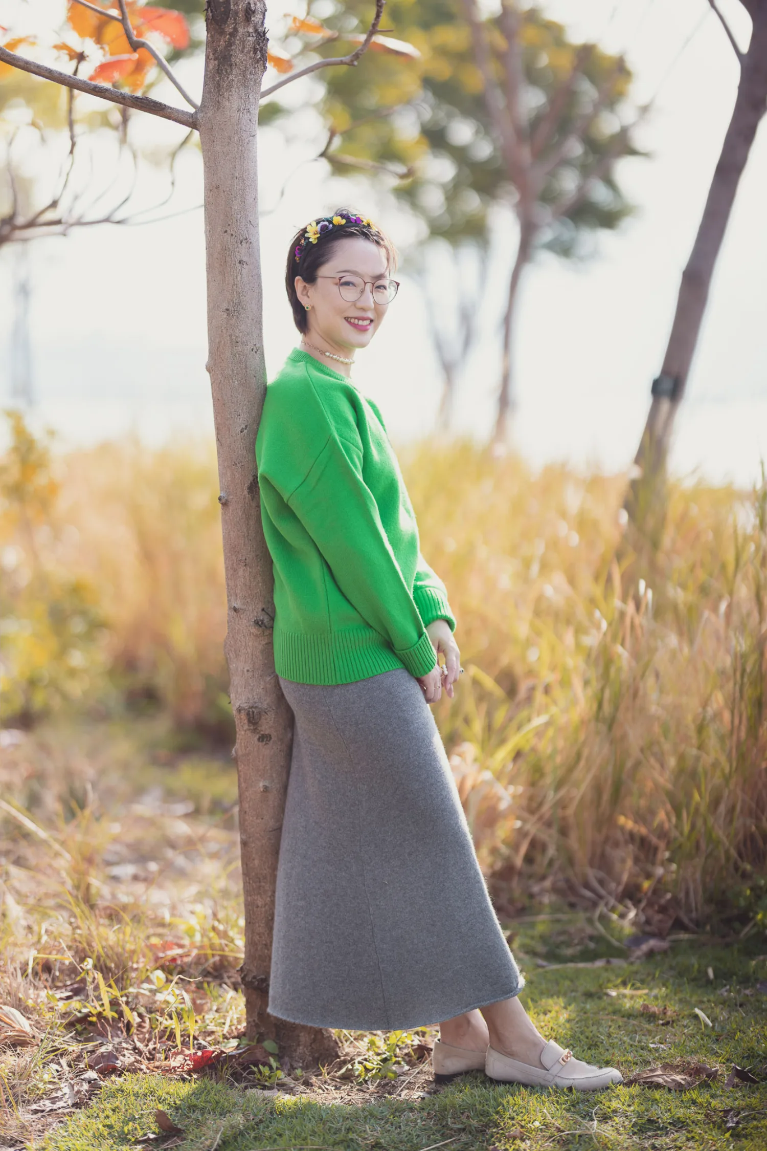 Bonnie leaning against a tree trunk at West Kowloon in her green sweater and floral headband, golden grasses catching the afternoon light