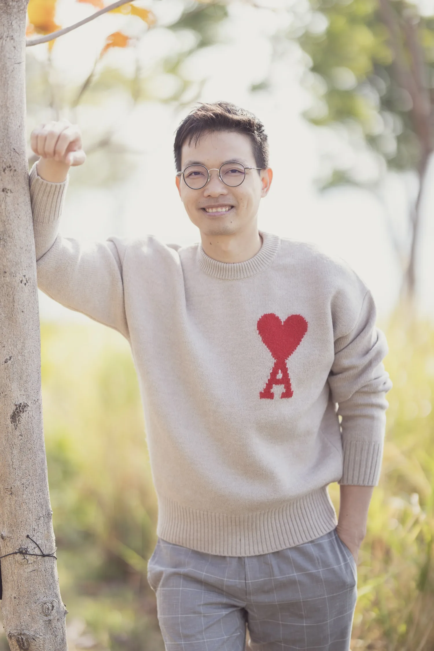 Vincent leaning casually against a tree at West Kowloon, wearing his Ami de Coeur heart sweater and round glasses