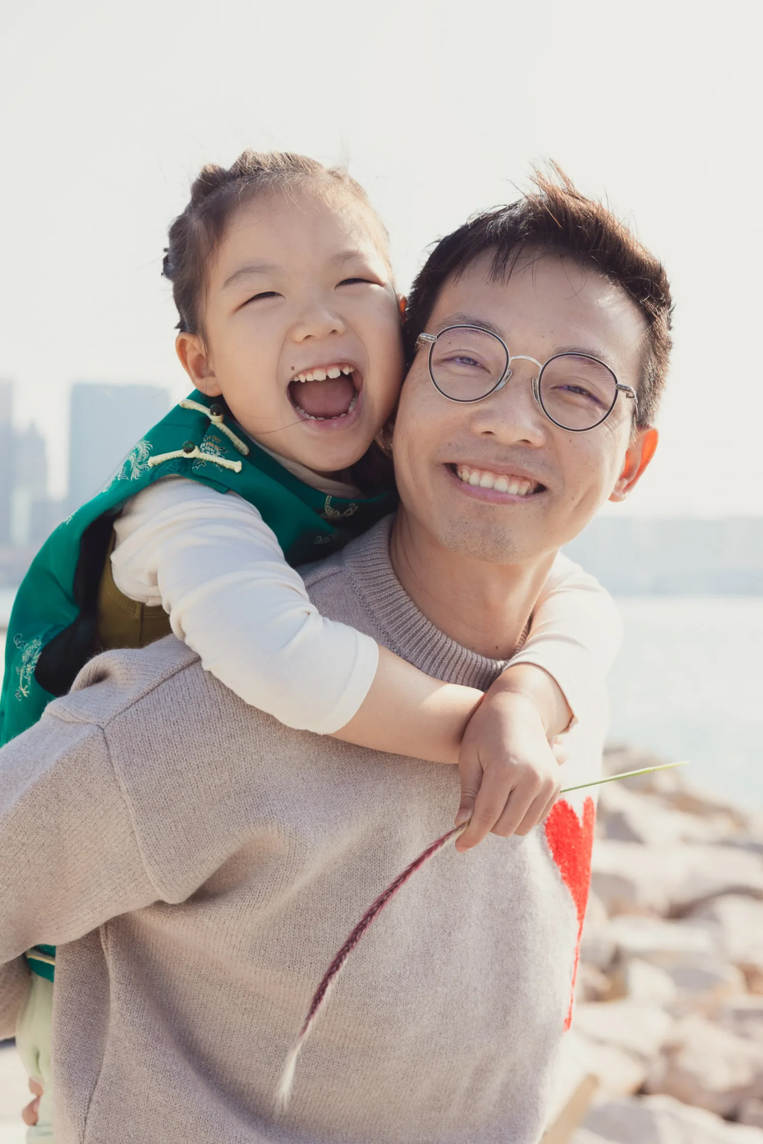 Emma piggybacking on Vincent by the rocky waterfront, both beaming — Hong Kong skyline and harbour behind them, warm golden light