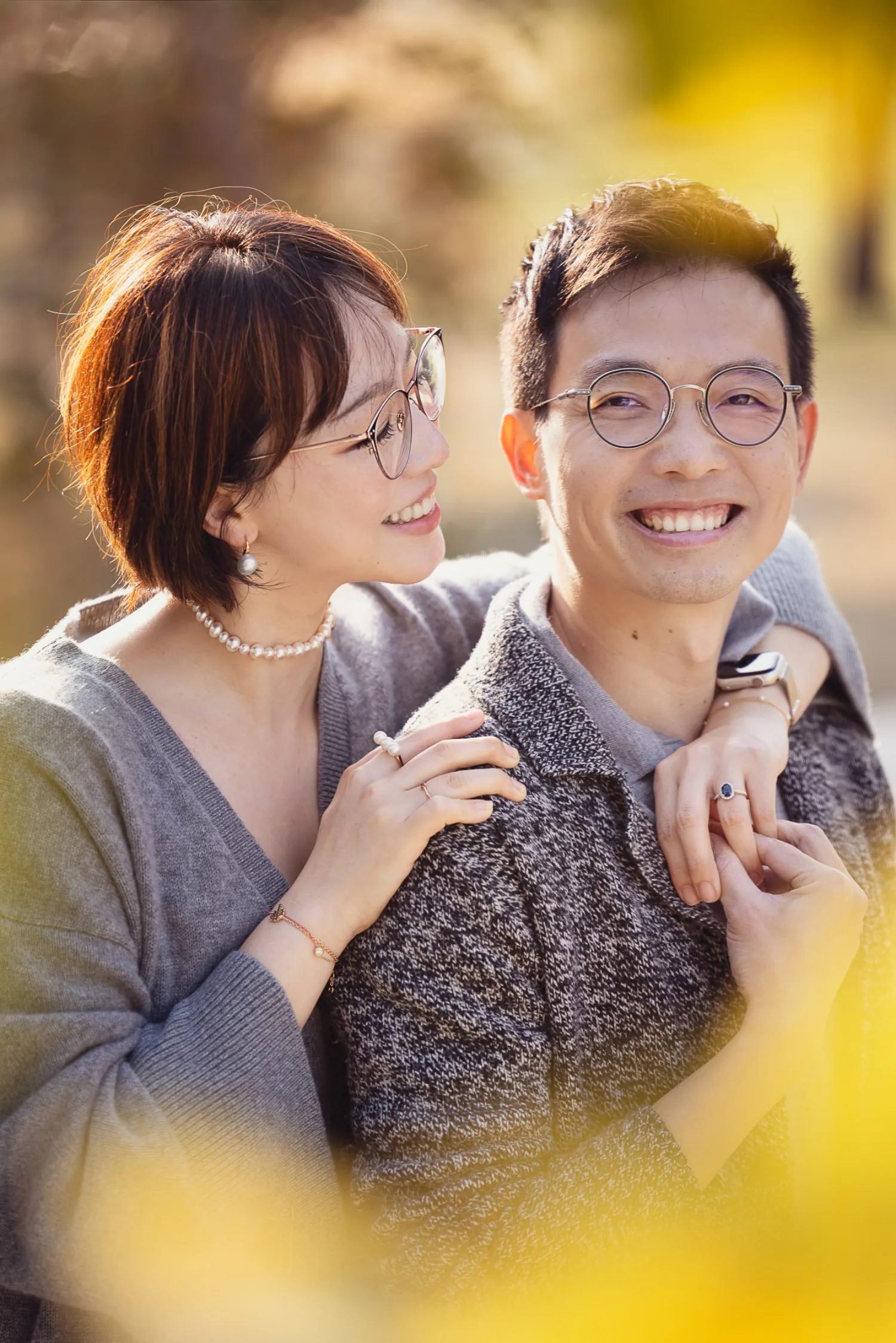 Bonnie and Vincent in a warm embrace at West Kowloon — Bonnie wrapping her arms around him from behind, both smiling in matching grey knitwear with golden backlight