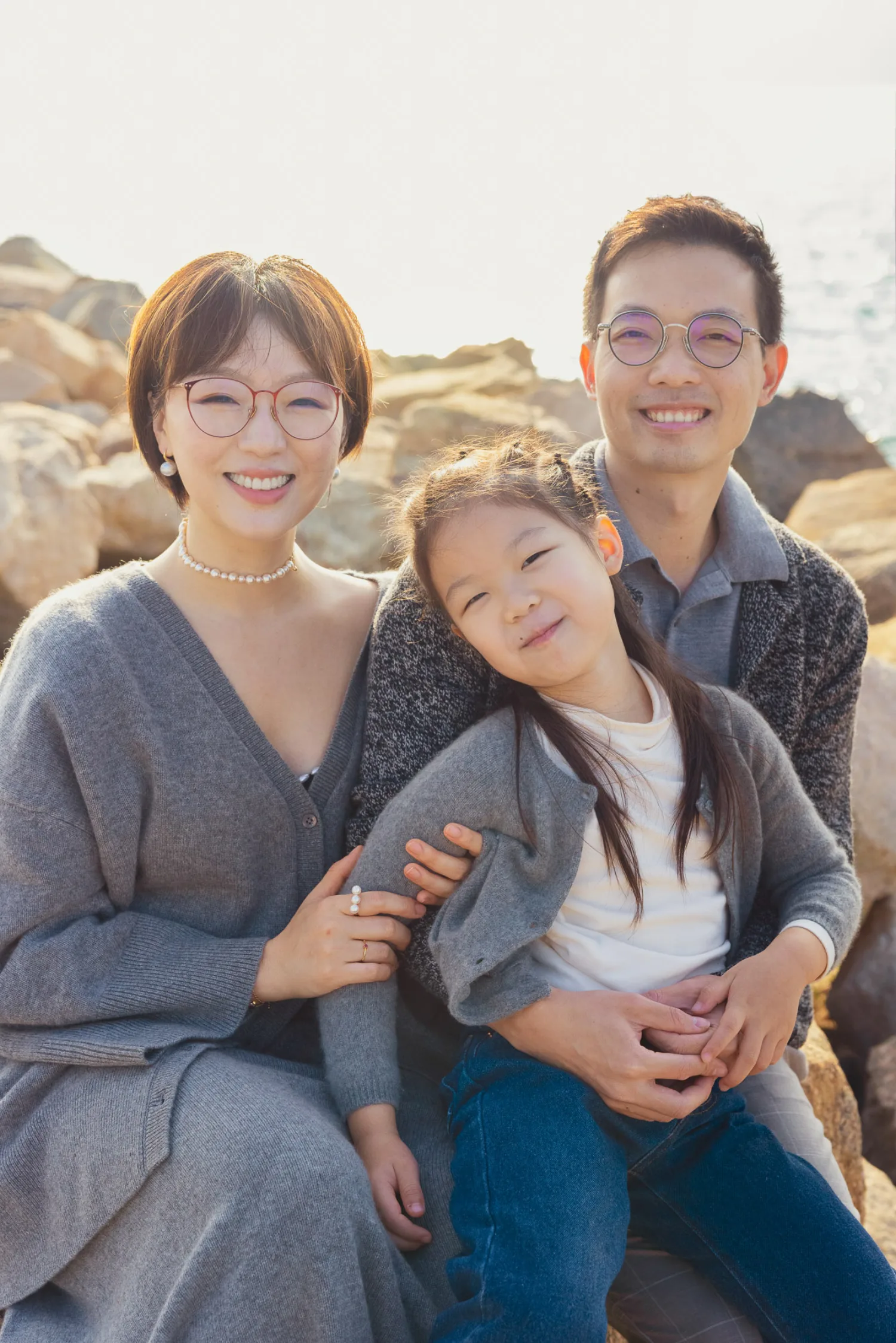 Family of three smiling at the camera on the rocky West Kowloon shoreline — Emma on Vincent's lap, Bonnie beside them, harbour behind
