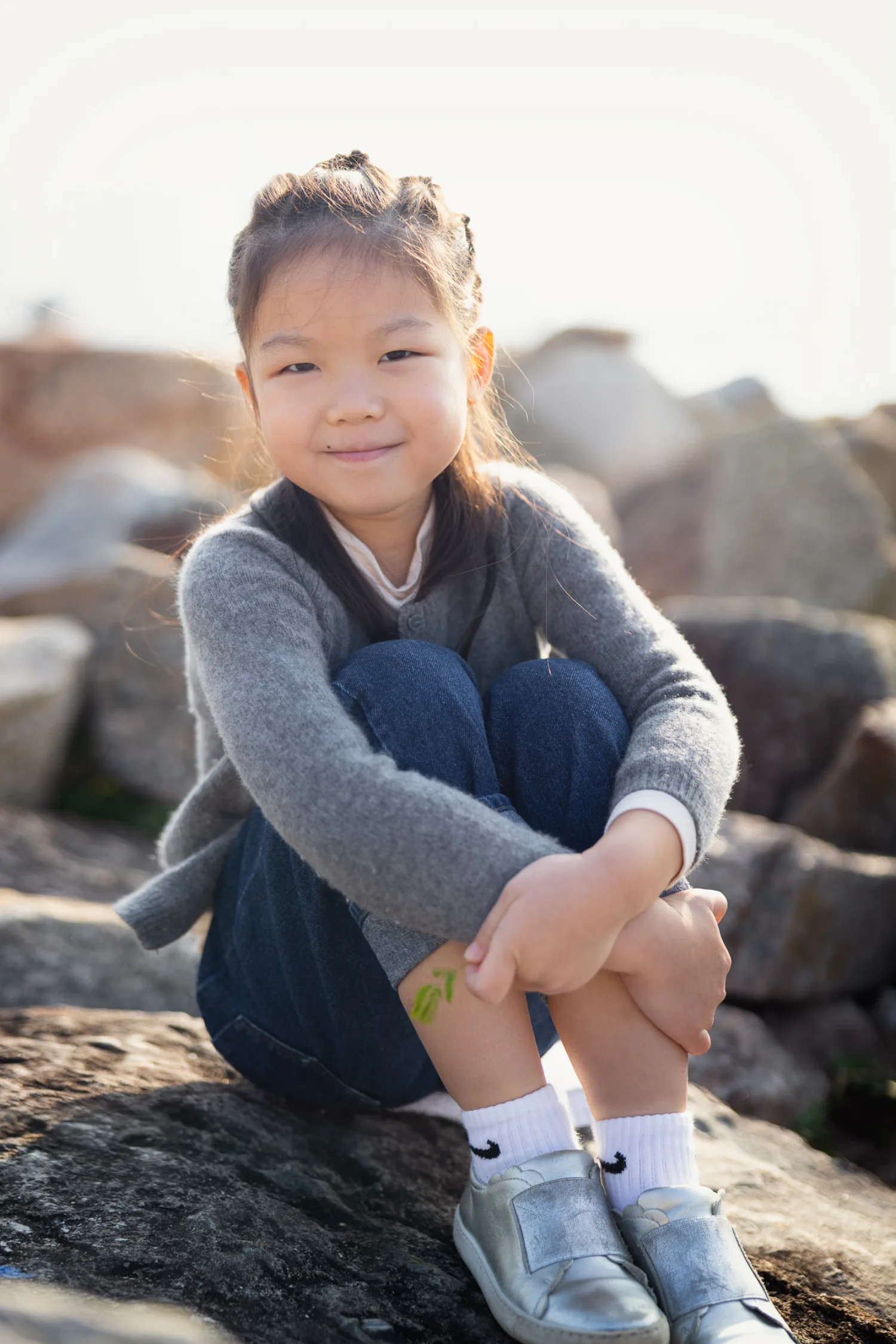Emma sitting on the rocks at West Kowloon, smiling at the camera in her grey cardigan and silver shoes with a little green drawing on her leg