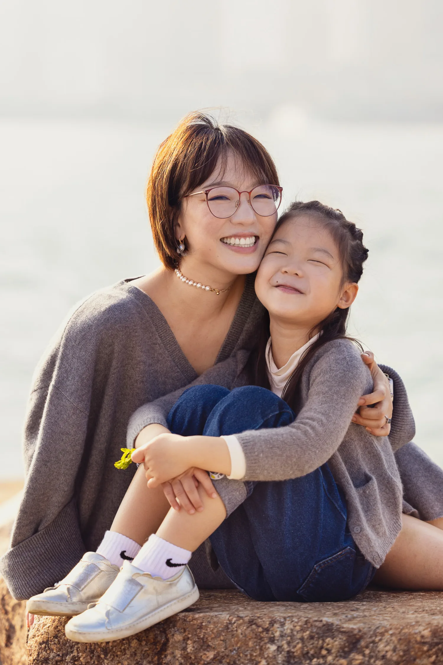 Bonnie and Emma sharing a joyful embrace on the stone surface at West Kowloon, both beaming — mum in grey sweater and glasses, daughter in grey cardigan and silver shoes